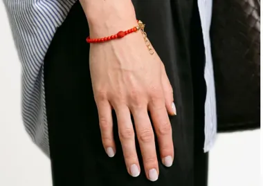 Dłoń z jasnym manicure i czerwoną bransoletką z koralików na nadgarstku.