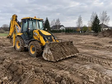 Żółta koparko-ładowarka JCB z pełną łyżką ziemi na placu budowy obok domów.