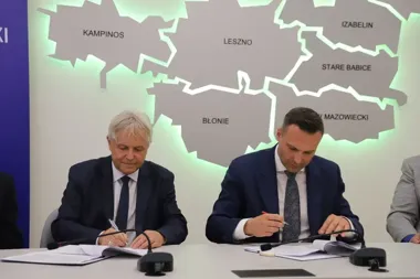 Dwaj mężczyźni w garniturach podpisują dokumenty przy stole, w tle podświetlana mapa z nazwami gmin.