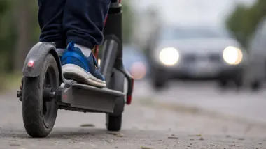 Zbliżenie na tylne koło hulajnogi elektrycznej, na której stoi osoba w niebieskich butach, z rozmytym samochodem z włączonymi światłami w tle.