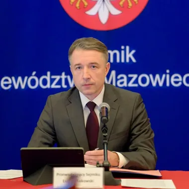 Mężczyzna w garniturze siedzi za stołem z mikrofonem podczas konferencji lub posiedzenia w siedzibie urzędu województwa mazowieckiego.