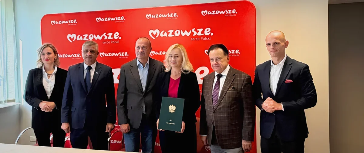 Grupa osób w strojach biznesowych pozuje do zdjęcia przed czerwoną ścianką z logotypem Mazowsze, jedna z kobiet trzyma zieloną teczkę z godłem.